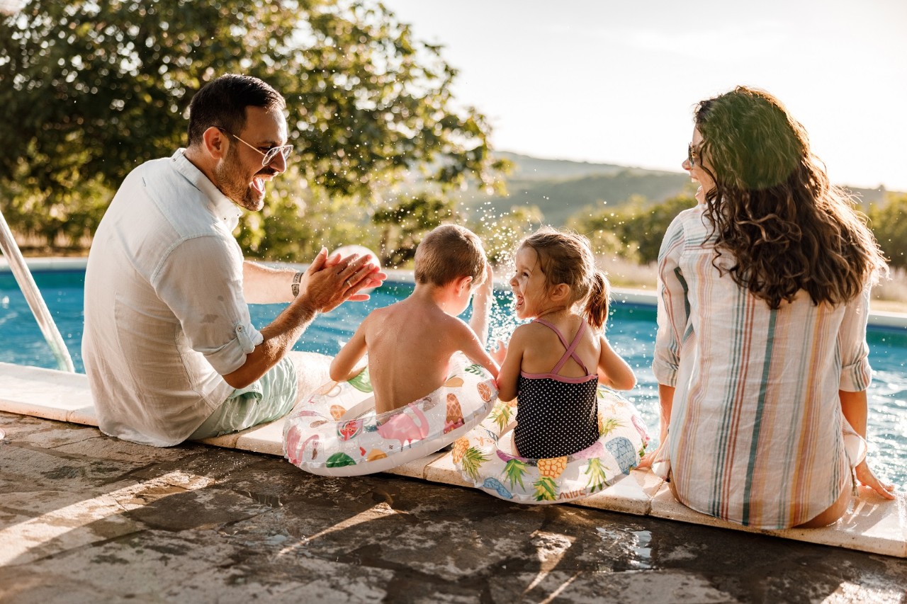 Happy parents and their small kids talking while relaxing by the pool at the backyard. Copy space.
