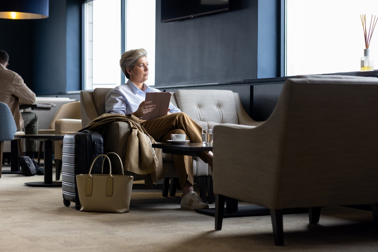 A full shot of a mature female adult sitting in an airport VIP lounge. She is holding a digital tablet sitting next to her carry-on luggage waiting for her gate to be called. The airport is located in the North East of England.

Videos are available similar to this scenario.
