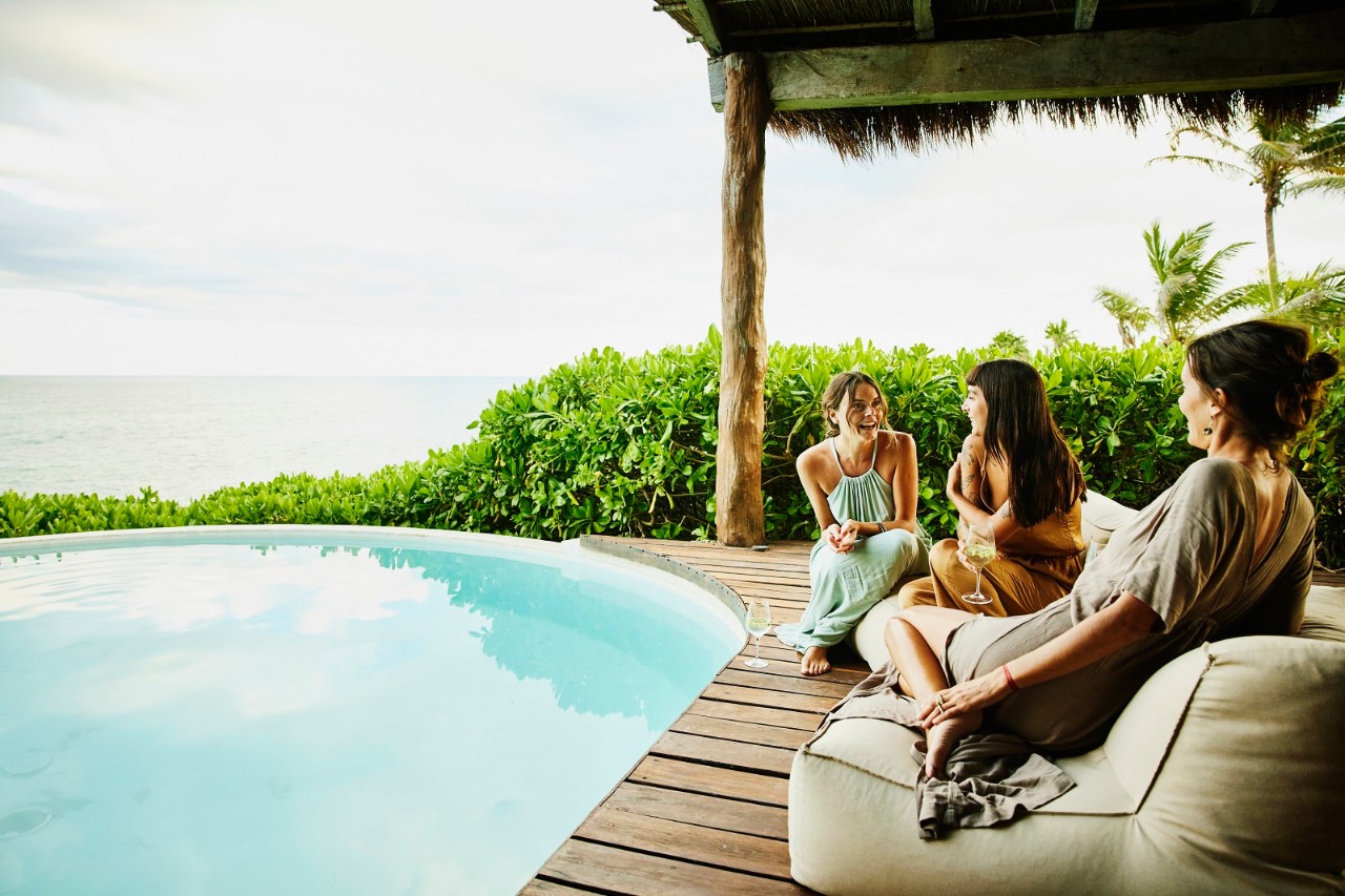 Wide shot of smiling female friends hanging out on deck of luxury suite at tropical resort
