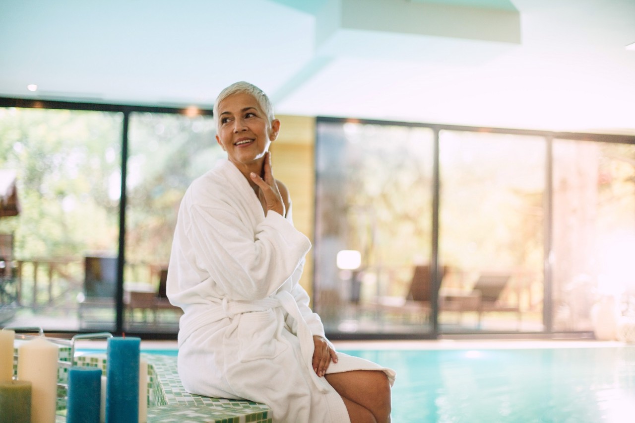 Mature woman is relaxing by the swimming pool at the spa. She is wearing a cozy bathrobe.