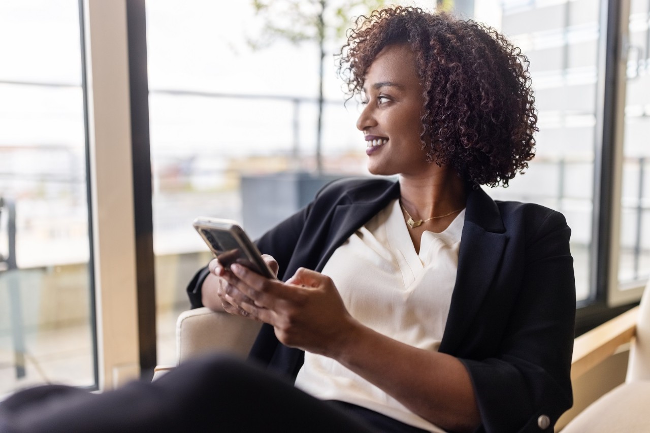 Happy african woman with curly hair sitting in hotel room with her mobile phone. Woman on business trip relaxing in her hotel room looking outside from the window and smiling.