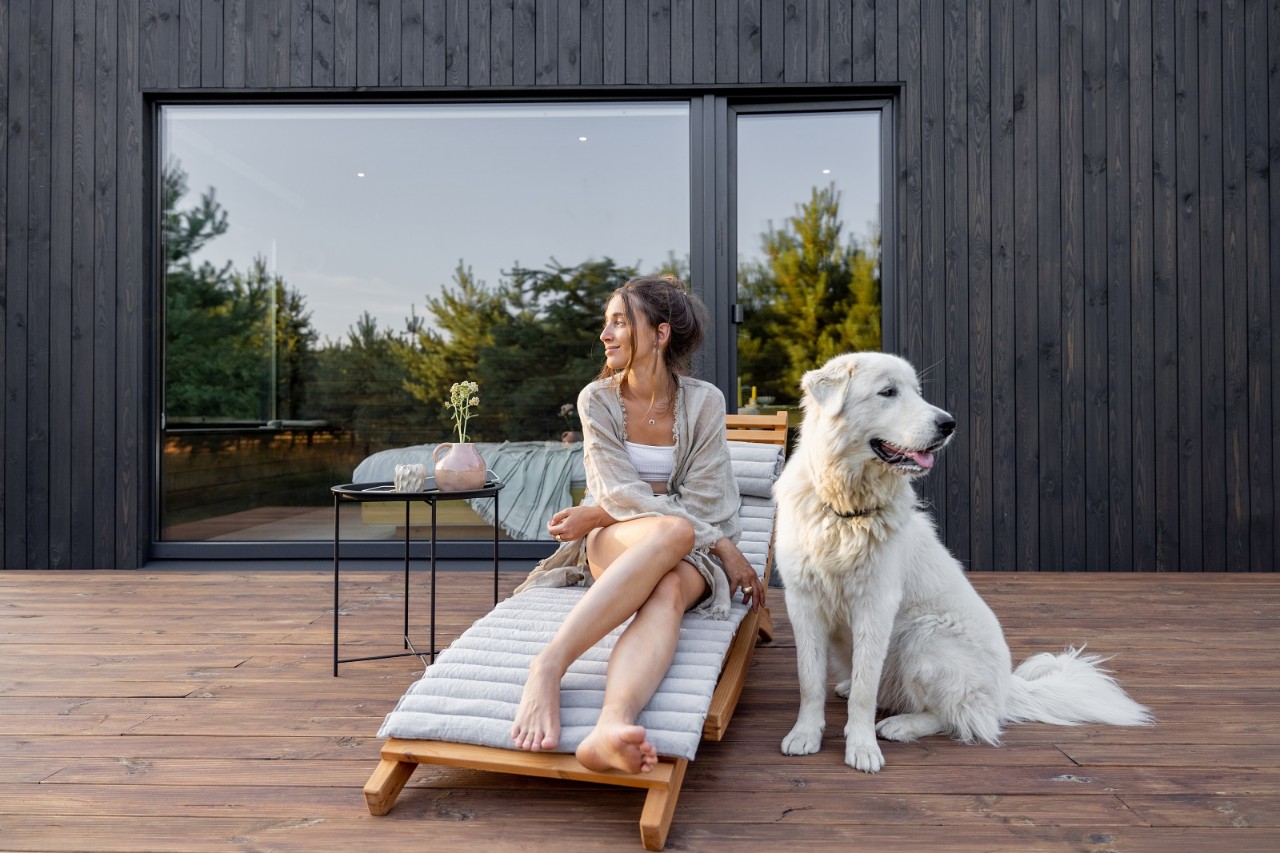 Woman enjoys the nature while sits on sunbed on wooden terrace at the modern hotel resort with panoramic windows near pine forest whith big white dog. Concept of solitude and recreation on nature