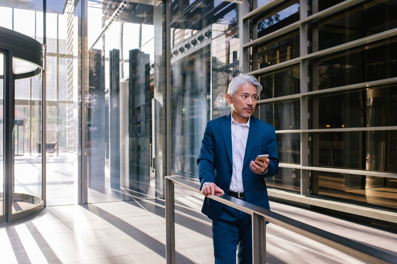 Businessman using his cell phone standing outdoors. Japanese man using his smartphone against an office building in background.
