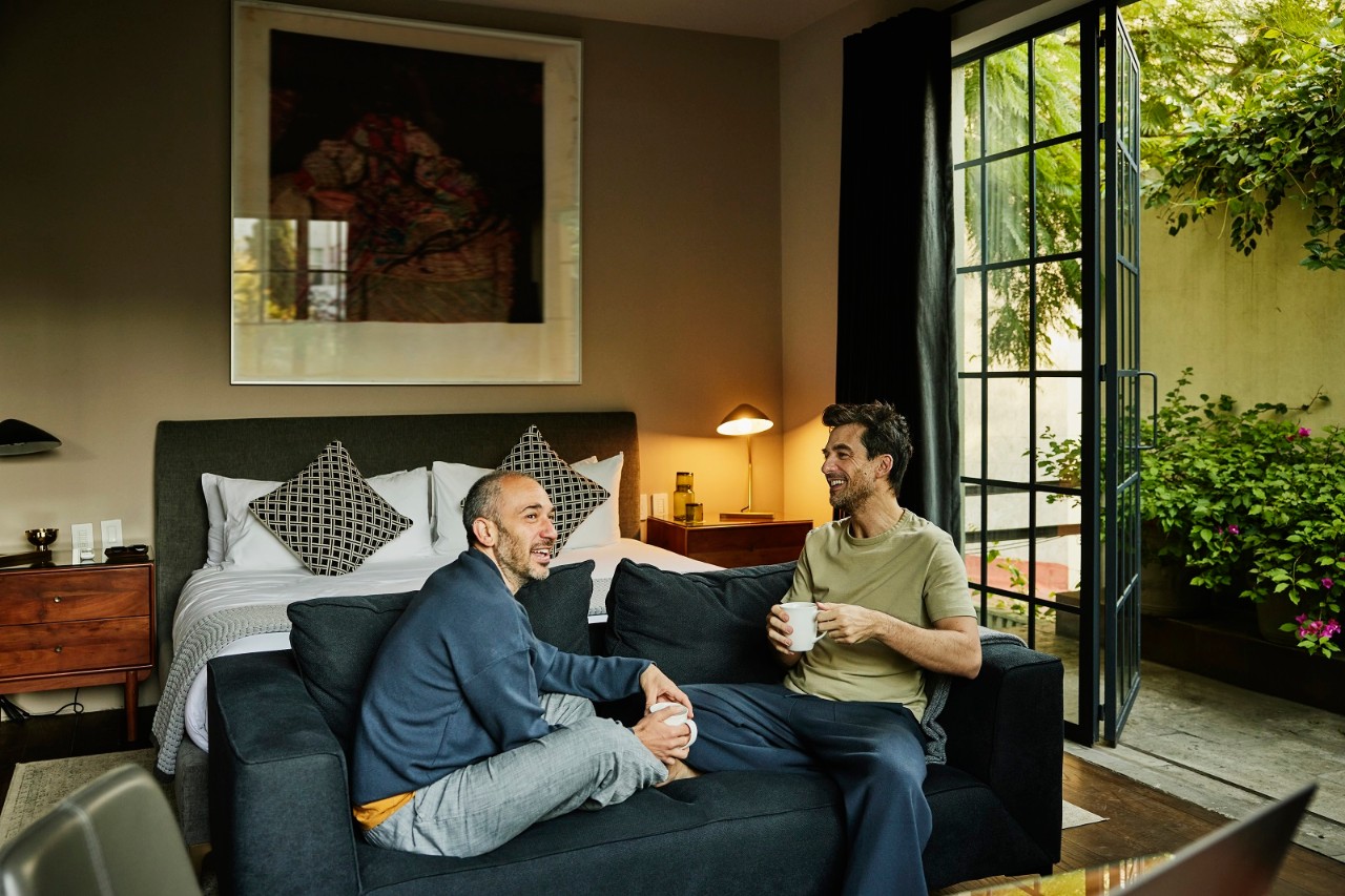Wide shot of smiling couple sharing morning coffee while relaxing in bedroom of boutique hotel suite during vacation