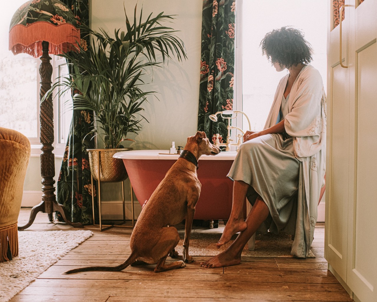 Serene image of an elegant domestic space with luxurious furniture. A woman perches on the side of a red roll top bath, wearing a silk robe. She waits for the bathtub to fill as her young lurcher watches on with curiosity.pours through the window, backlighting the tranquil scene, giving it a dreamy vibe.